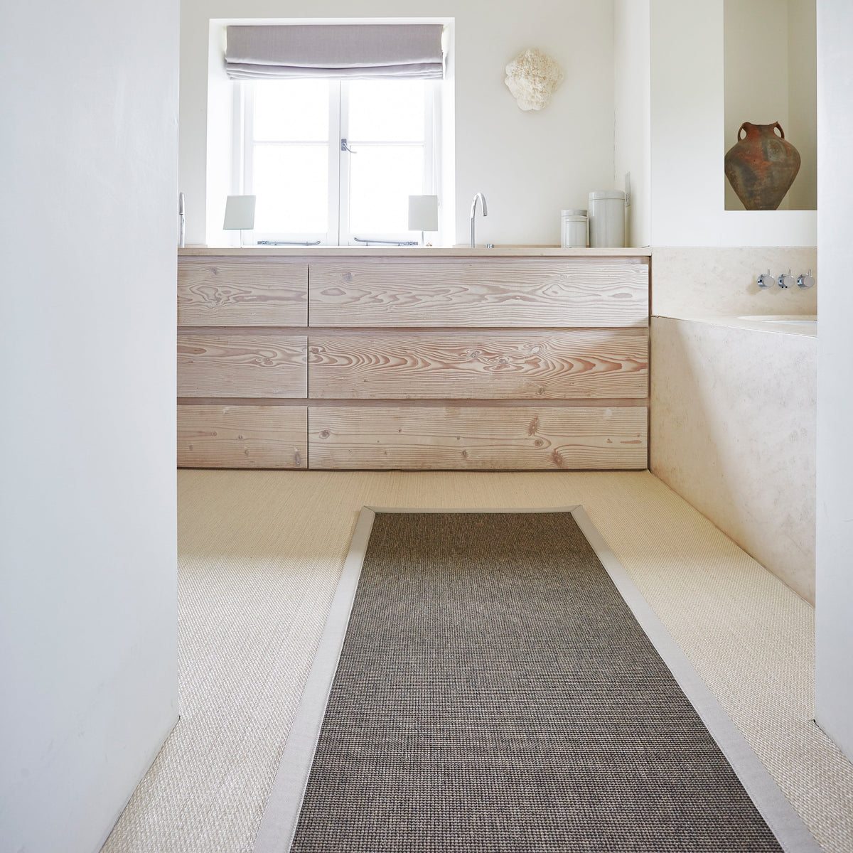 A modern bathroom interior featuring a light wood dresser against a window, a textured gray runner rug on a cream-colored floor, and a recessed niche with a ceramic vase.